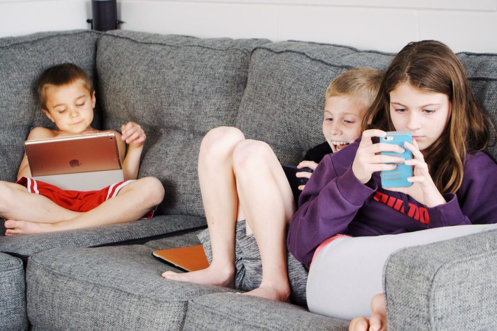Positive barefoot children in casual wear resting together on cozy couch and browsing tablets and smartphones